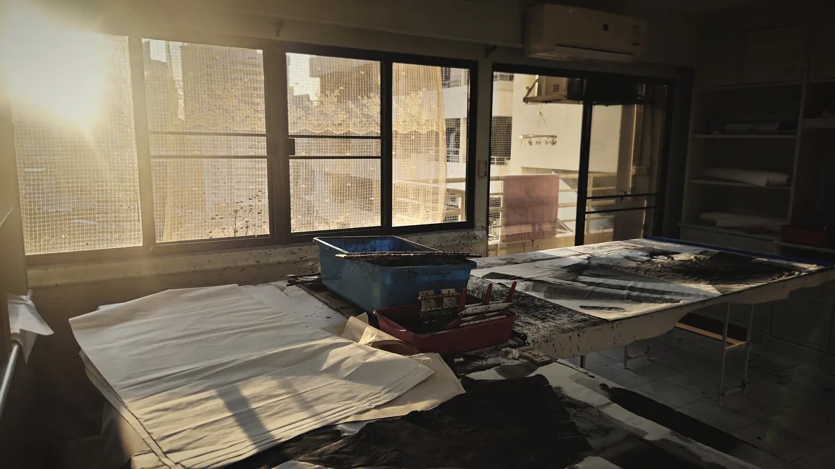 The studio at sunrise: a long felt-covered table under a wall of windows, blank sheets stacked at one end and painted sheets drying at the other